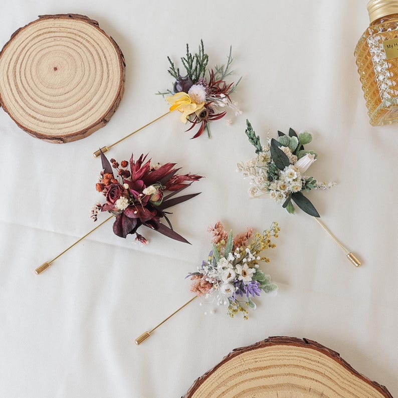 Puede incluir: Cuatro prendedores florales con tallos dorados y arreglos florales decorativos en varios colores, incluyendo burdeos, amarillo y blanco. Los prendedores est&aacute;n dispuestos sobre una superficie blanca, con rodajas de madera y un frasco de perfume en el fondo.
