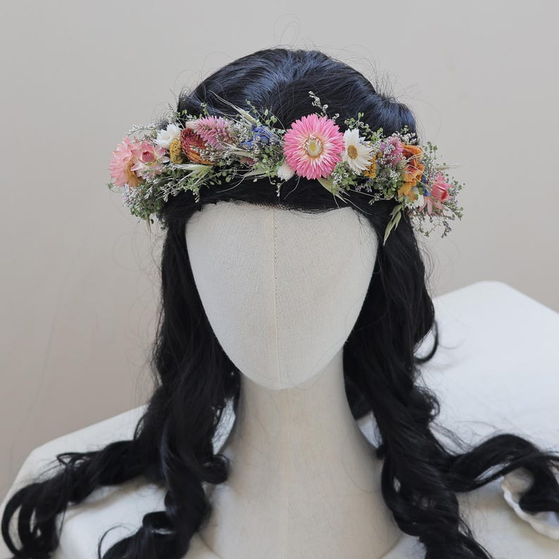 May include: A floral crown featuring a variety of dried flowers in shades of pink, white, yellow, and orange, accented with green foliage. The crown is set against a backdrop of black hair and a neutral background, creating a delicate and natural aesthetic.
