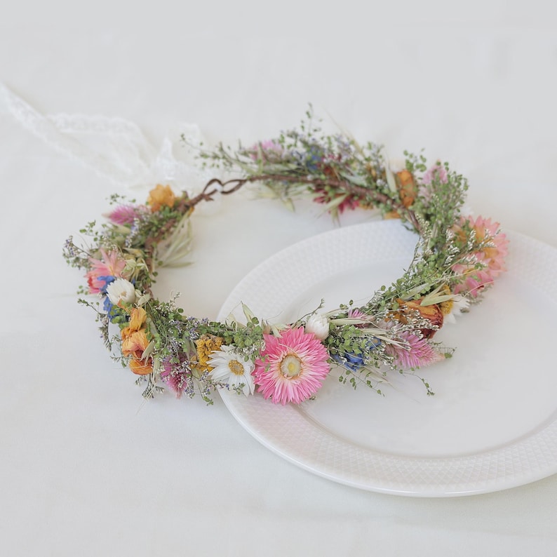 May include: A floral crown featuring a variety of dried flowers in shades of pink, yellow, orange, and white, accented with green foliage. The crown is tied with a white lace ribbon, perfect for a wedding or special occasion.