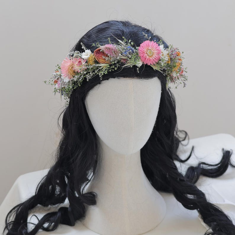 May include: A floral crown featuring a variety of dried flowers in shades of pink, white, and yellow, accented with green foliage. The crown is displayed on a mannequin head with black wavy hair, perfect for weddings or special events.