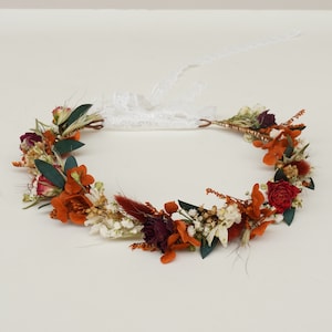 May include: A floral crown with orange, red, and white dried flowers and green leaves. The crown is made with a copper wire base and has a white lace ribbon.