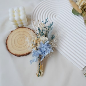 May include: A small bouquet of dried flowers with blue hydrangeas, white and cream flowers, and green foliage. The bouquet is tied with twine and is ready for a wedding or special occasion.