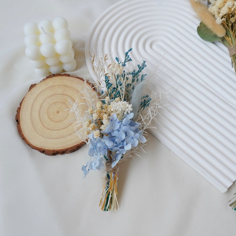 May include: A small bouquet of dried flowers with blue hydrangeas, white and cream flowers, and green foliage. The bouquet is tied with twine and is ready for a wedding or special occasion.