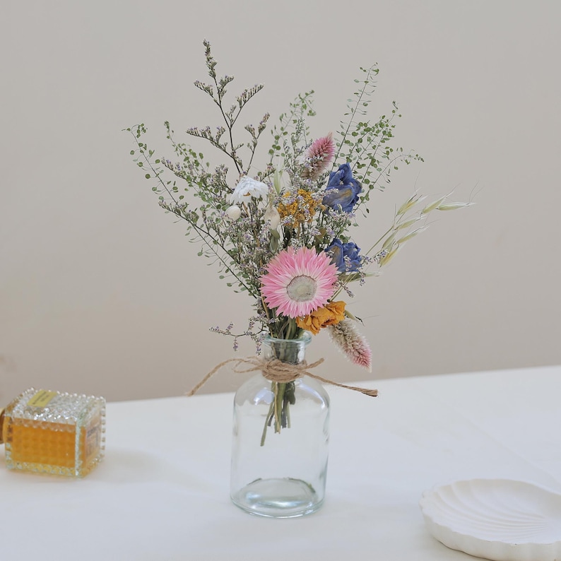 May include: A small, clear glass vase holds a bouquet of dried flowers. The arrangement includes pink, blue, yellow, and white blooms, with green foliage. The vase is tied with twine. A small, decorative box is visible to the left.