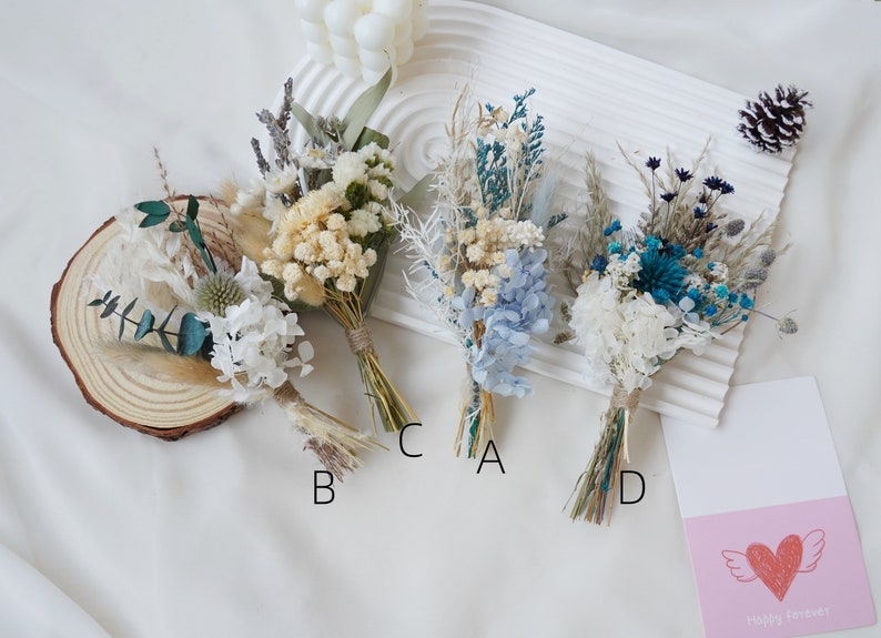 May include: Four small bouquets of dried flowers, each tied with twine. The bouquets are arranged in a row on a white surface. The bouquets are labeled A, B, C, and D. The bouquets are made up of dried flowers in shades of white, blue, and green.