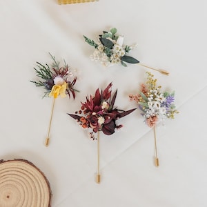 Puede incluir: Cuatro prendedores florales con alfileres dorados. Los prendedores presentan una variedad de flores secas en tonos rojos, amarillos, blancos y morados. Los prendedores est&aacute;n dispuestos sobre una superficie blanca.
