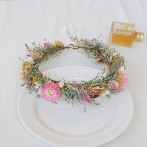 May include: A vibrant floral crown featuring a variety of dried flowers in shades of pink, yellow, and blue, accented with green foliage. The crown is displayed on a white plate, perfect for a wedding or special occasion.