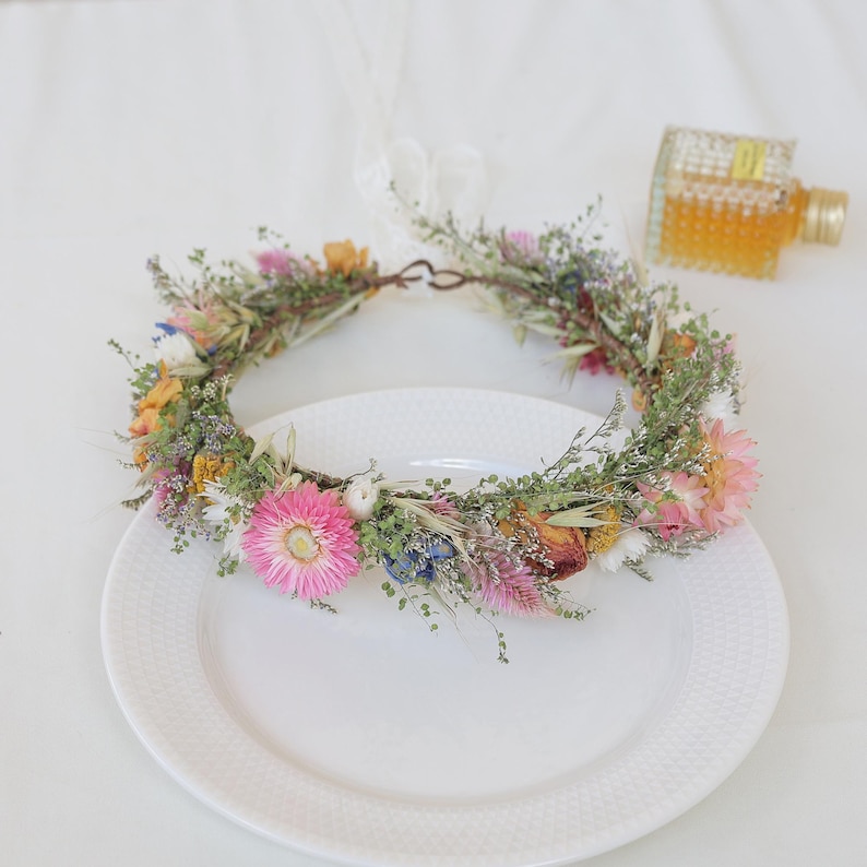 May include: A vibrant floral crown featuring a variety of dried flowers in shades of pink, yellow, and blue, accented with green foliage. The crown is displayed on a white plate, perfect for a wedding or special occasion.