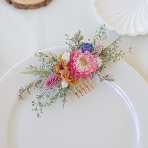 May include: A floral hair comb featuring a vibrant arrangement of dried flowers in shades of pink, orange, blue, and white. The flowers are attached to a gold-colored comb, perfect for adding a touch of nature-inspired elegance to any hairstyle.