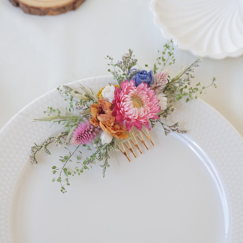 May include: A floral hair comb featuring a vibrant arrangement of dried flowers in shades of pink, orange, blue, and white. The flowers are attached to a gold-colored comb, perfect for adding a touch of nature-inspired elegance to any hairstyle.