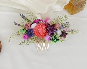 May include: A floral hair comb featuring a vibrant arrangement of dried flowers in shades of pink, purple, and green. The flowers are attached to a gold-colored comb. The comb is designed for bridal or special occasion hairstyles.