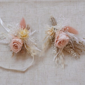 May include: Two dried flower arrangements with pink roses, white and beige dried grasses, and a white ribbon. The arrangements are on a beige fabric background.