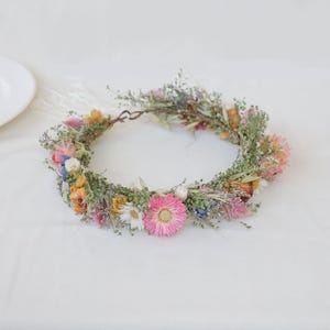 May include: A floral crown featuring a variety of dried flowers in shades of pink, yellow, white, and blue. The crown is made of a woven base and is adorned with delicate greenery, creating a natural and whimsical look.