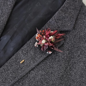 Puede incluir: Una blazer de tweed gris oscuro con un prendedor floral. El prendedor presenta flores y follaje secos de color burdeos, blanco y naranja. Un alfiler de solapa dorado asegura el arreglo floral a la chaqueta.