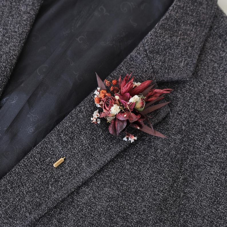 Puede incluir: Una blazer de tweed gris oscuro con un prendedor floral. El prendedor presenta flores y follaje secos de color burdeos, blanco y naranja. Un alfiler de solapa dorado asegura el arreglo floral a la chaqueta.