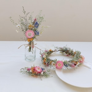 May include: A collection of dried floral arrangements. Includes a bouquet in a glass bottle, a floral crown, and a hair comb. The flowers feature pink, blue, yellow, and white blooms with green foliage. The items are arranged on a white surface.
