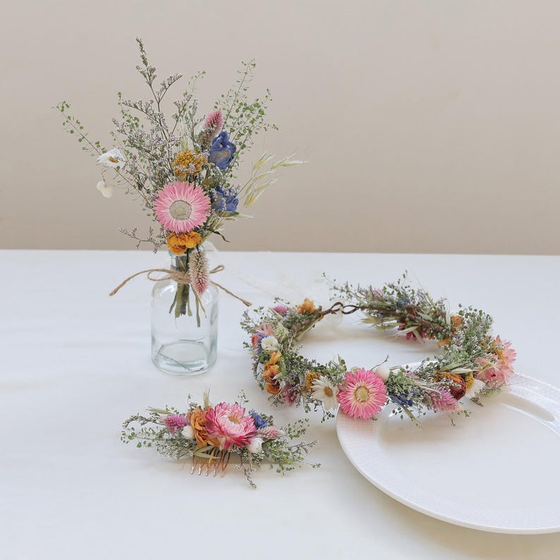 May include: A collection of dried floral arrangements. Includes a bouquet in a glass bottle, a floral crown, and a hair comb. The flowers feature pink, blue, yellow, and white blooms with green foliage. The items are arranged on a white surface.