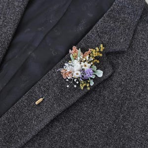 Puede incluir: Un prendedor floral con flores secas en tonos blancos, morados, amarillos y melocot&oacute;n. El prendedor est&aacute; sujeto a una blazer de tweed gris oscuro con un alfiler dorado.