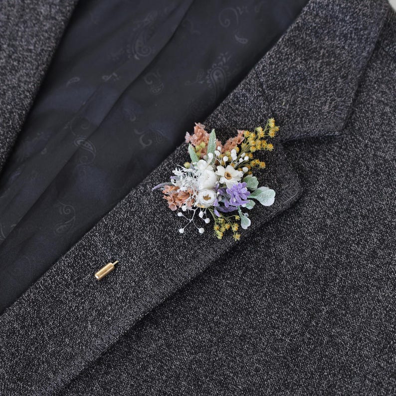 Puede incluir: Un prendedor floral con flores secas en tonos blancos, morados, amarillos y melocot&oacute;n. El prendedor est&aacute; sujeto a una blazer de tweed gris oscuro con un alfiler dorado.
