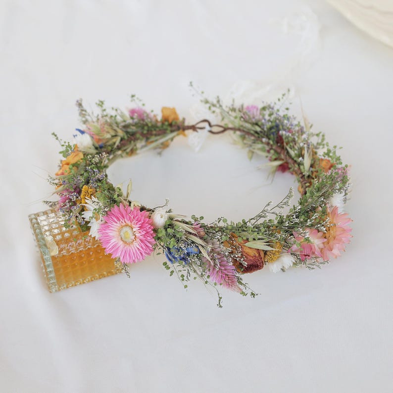 May include: A floral crown featuring a variety of dried flowers in shades of pink, yellow, blue, and green. The crown is arranged in a circular shape, with a metal wire base. A small, decorative glass piece is visible.