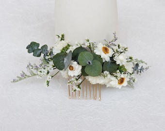 May include: A gold hair comb adorned with white flowers and green leaves. The comb is perfect for adding a touch of elegance to any hairstyle.