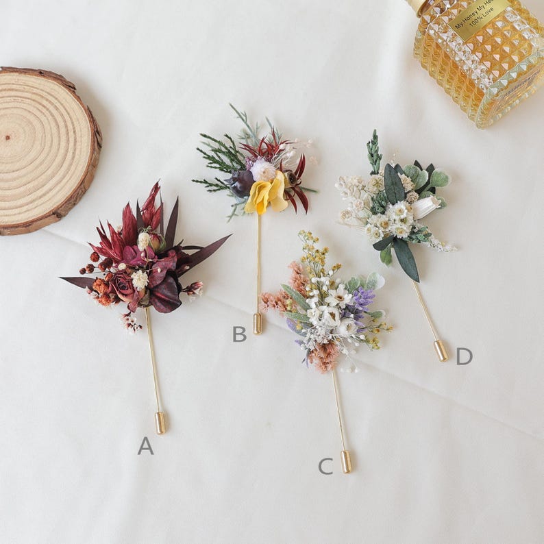 Puede incluir: Cuatro prendedores florales con tallos dorados y arreglos florales decorativos. Los prendedores presentan varios colores, incluyendo burdeos, amarillo, blanco y morado. Los prendedores est&aacute;n dispuestos sobre una superficie blanca.