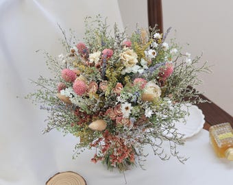 May include: A bouquet of dried flowers featuring pink globe amaranth, cream-colored roses, purple lavender, yellow filler flowers, and green foliage. The arrangement is tied at the base and includes various textures of dried grasses and small white blossoms.