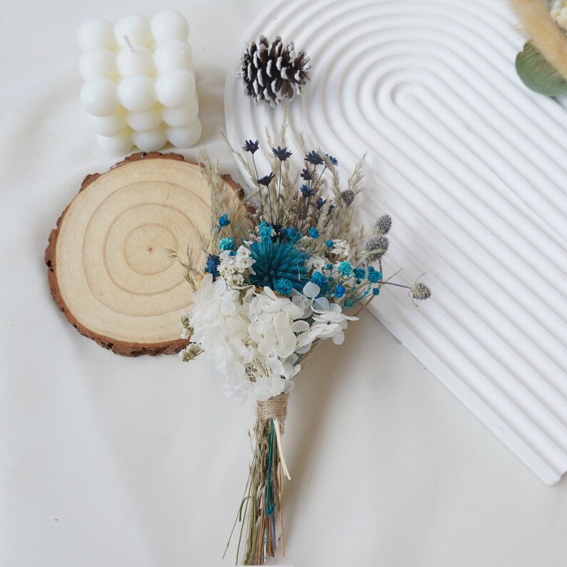 May include: A small bouquet of dried flowers in shades of blue and white, tied with twine. The bouquet includes white hydrangeas, blue and white baby's breath, and dried grasses.