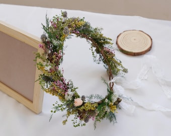 May include: A floral crown made with dried flowers and greenery. The crown is a circle with a white lace ribbon attached. The crown is made with a variety of flowers, including pink, yellow, and white flowers.