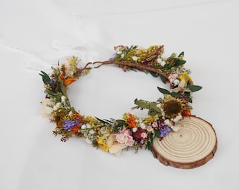 May include: A circular floral crown composed of dried flowers in shades of yellow, orange, purple, pink, and green, featuring small white blossoms and green foliage. The crown includes thin white lace ribbons and rests near a round wooden slice.