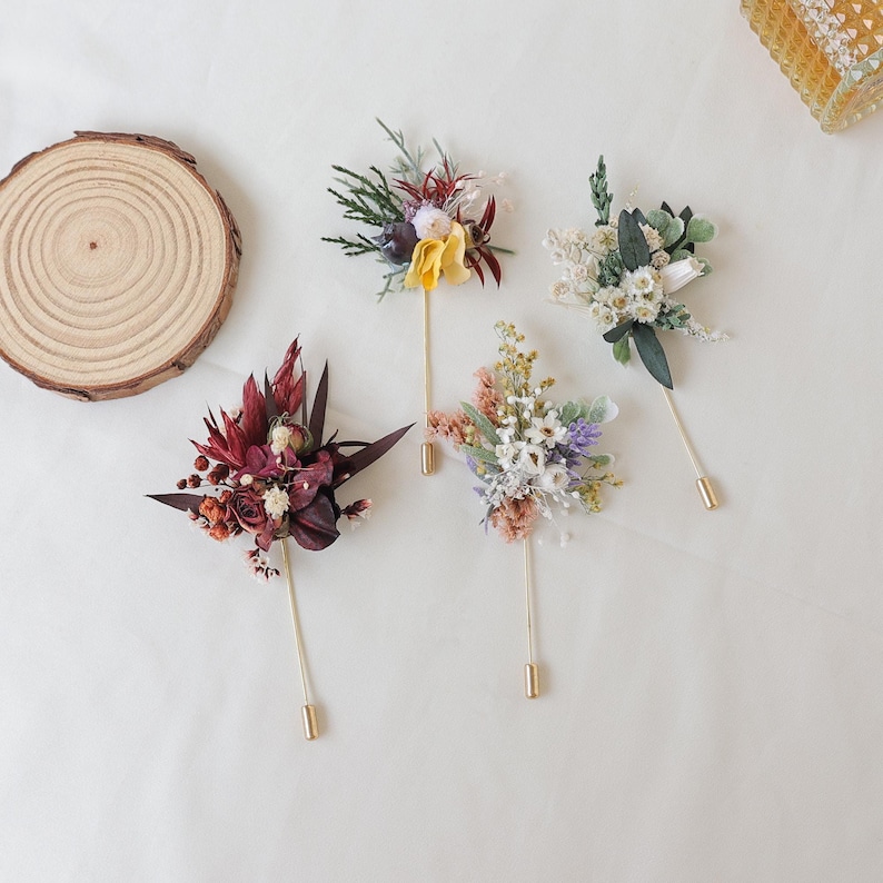 Puede incluir: Cuatro boutonnieres florales con alfileres dorados. Las flores son de varios colores, incluyendo burdeos, amarillo, blanco y lavanda. Una rodaja de madera est&aacute; en la esquina superior izquierda. Los boutonnieres est&aacute;n dispuestos sobre una superficie blanca.