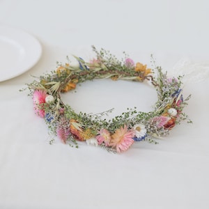 May include: A floral crown featuring a variety of dried flowers in shades of pink, yellow, blue, and white. The crown is adorned with greenery and has a natural, rustic appearance. It is tied with a white ribbon.