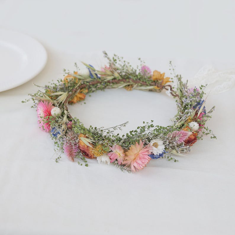 May include: A floral crown featuring a variety of dried flowers in shades of pink, yellow, blue, and white. The crown is adorned with greenery and has a natural, rustic appearance. It is tied with a white ribbon.