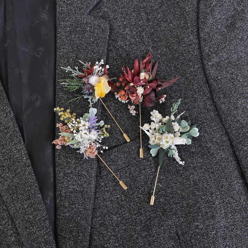 Puede incluir: Tres prendedores florales con tallos dorados est&aacute;n prendidos a una americana gris. Los prendedores presentan flores secas en tonos rojos, amarillos, blancos y morados. La americana es de tejido texturizado.