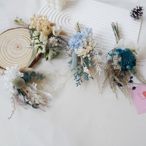 May include: Four small bouquets of dried flowers in various shades of blue, white, and cream. The bouquets are arranged on a white surface with a wooden round slice and a pink card with a heart and the text "Happy Birthday".