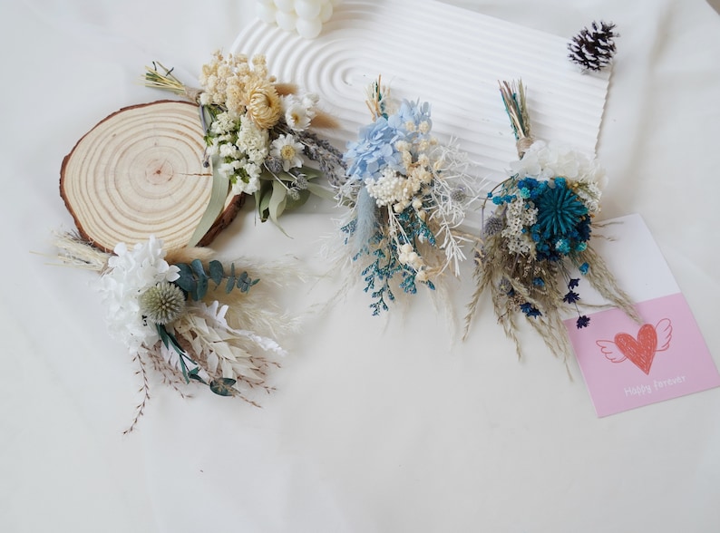May include: Four small bouquets of dried flowers in various shades of blue, white, and cream. The bouquets are arranged on a white surface with a wooden round slice and a pink card with a heart and the text "Happy Birthday".