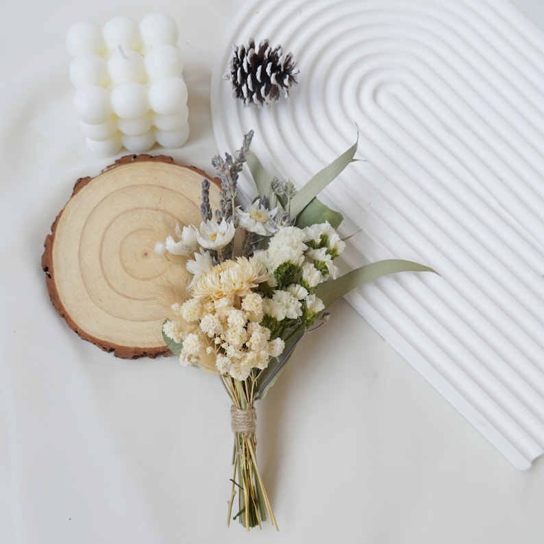 May include: A small bouquet of dried flowers in shades of white and cream, tied with twine. The bouquet is arranged on a wooden slice and includes dried lavender, baby's breath, and other delicate flowers.