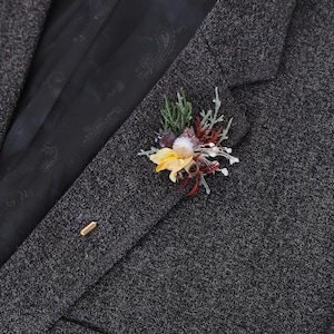 Puede incluir: Un blazer de tweed gris con un prendedor floral. El prendedor presenta flores amarillas, rojas y blancas con follaje verde. Tambi&eacute;n se ve un prendedor dorado.