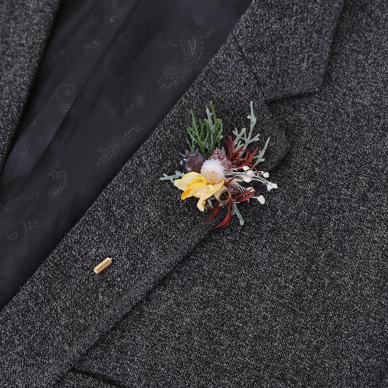 Puede incluir: Un blazer de tweed gris con un prendedor floral. El prendedor presenta flores amarillas, rojas y blancas con follaje verde. Tambi&eacute;n se ve un prendedor dorado.