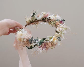 May include: A delicate floral crown made with dried flowers in shades of white, pink, and green. The crown is adorned with a blush pink ribbon.