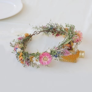 May include: A floral crown made of dried flowers in shades of pink, yellow, white, and blue. The crown is adorned with greenery and rests next to a small, clear bottle with a gold cap.