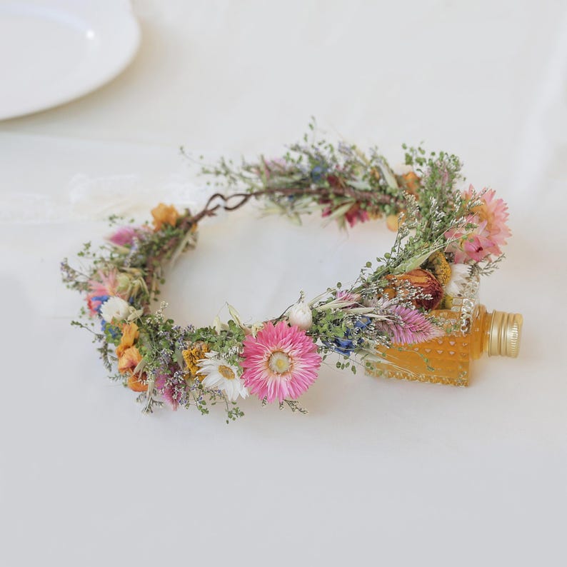 May include: A floral crown made of dried flowers in shades of pink, yellow, white, and blue. The crown is adorned with greenery and rests next to a small, clear bottle with a gold cap.