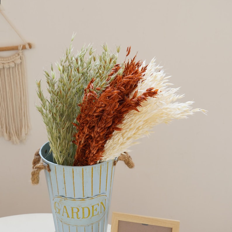 May include: A blue metal vase with a white stripe pattern and the word "GARDEN" printed on the front. The vase is filled with dried pampas grass in shades of white, brown, and green.