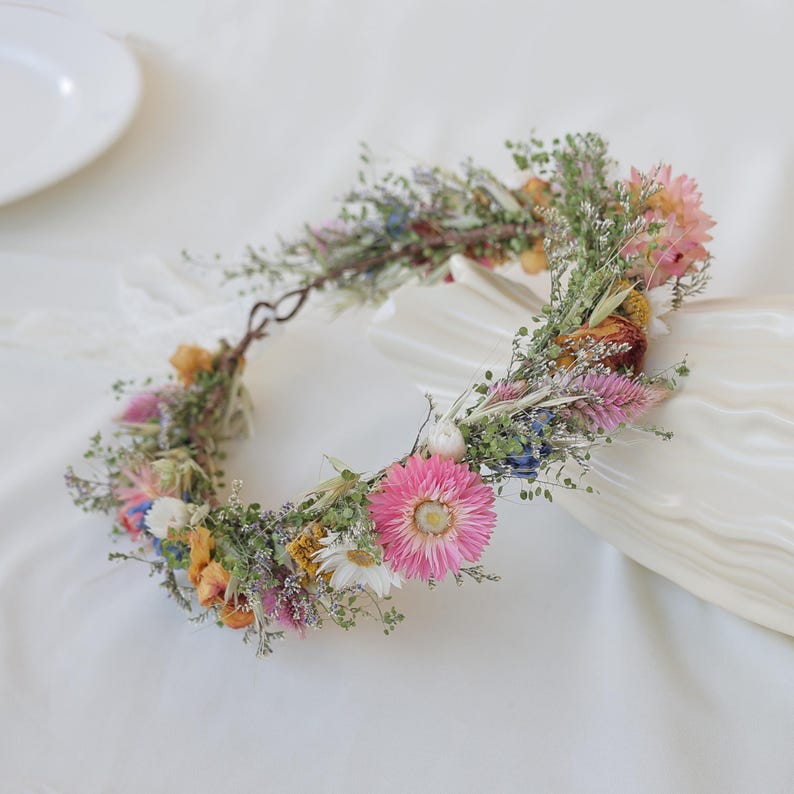 May include: A floral crown featuring a variety of dried flowers in shades of pink, yellow, blue, and white. The crown is made of a natural twig base and is adorned with delicate greenery, creating a rustic and elegant look.