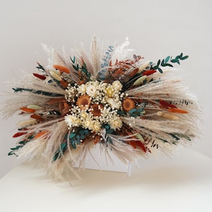 May include: A dried flower arrangement in a white box. The arrangement features a variety of dried grasses, pampas grass, and flowers in shades of beige, brown, and green.