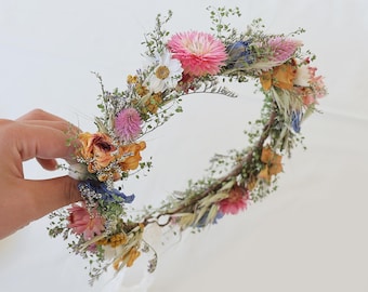May include: A handmade floral crown featuring a variety of dried flowers in shades of pink, orange, blue, and white. The crown is made with a natural vine base and delicate greenery, perfect for a bohemian or rustic aesthetic.