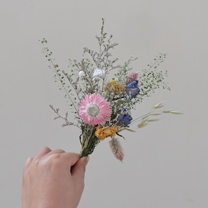 May include: A small bouquet of dried flowers featuring a large pink flower at the center. The bouquet includes blue, yellow, and white flowers, with green stems and sprigs. The bouquet is held in a hand against a neutral background.