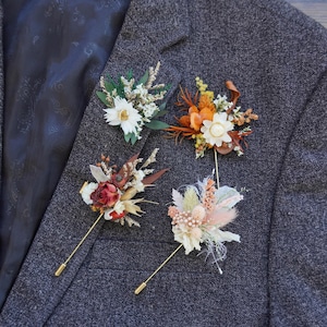 May include: Four floral boutonnieres with gold pins are displayed on a grey tweed blazer. The boutonnieres feature dried flowers in white, orange, red, and pink hues, with green accents. The blazer's interior lining has a dark paisley pattern.