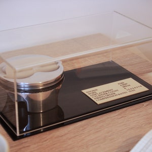May include: A clear acrylic display case with a black base, showcasing a silver engine piston. The base has a gold plaque with the text "ENGINE, BORE: 4.000", "DISPLACEMENT: 350", "CYLINDER COUNT: 8", "FIRING ORDER: 1-5-4-2-6-3-7-8", "COMPRESSION RATIO: 11.0:1".
