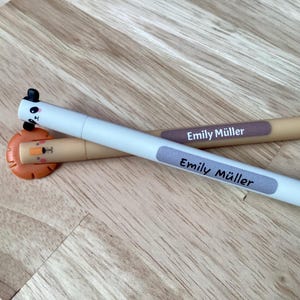 Könnte beinhalten: Zwei Stifte mit Tiermotiven. Einer ist weiß mit einem Pandakopf, der andere ist beige mit einem Löwenkopf. Beide Stifte tragen den Namen "Emily Müller". Sie liegen auf einer Holzoberfläche.